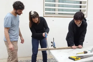 Une adulte utilise une scie à bois pour couper un tasseau maintenu par une autre adulte. Un animateur supervise.