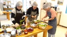Trois adultes sont dans une cuisine, avec un tablier et une charlotte sur la tête. Elles sélectionnent des bouts de légumes qu'il y a sur la table.