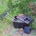 Photo d'une malle noire à moitié ouverte avec du materiel d'observation dedans, dessus et à côté : filet à papillon, épuisette, livres, jumelles, boites-loupes, loupes...