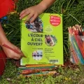 On voit des mains d'enfants tenir de face le livre l'Ecole à ciel ouvert tout en manipulant des crayons de couleurs. 